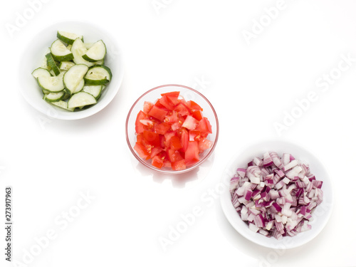 Sliced tomato in a transparent bowl, red onions and cucumbers in a white bowls on white background