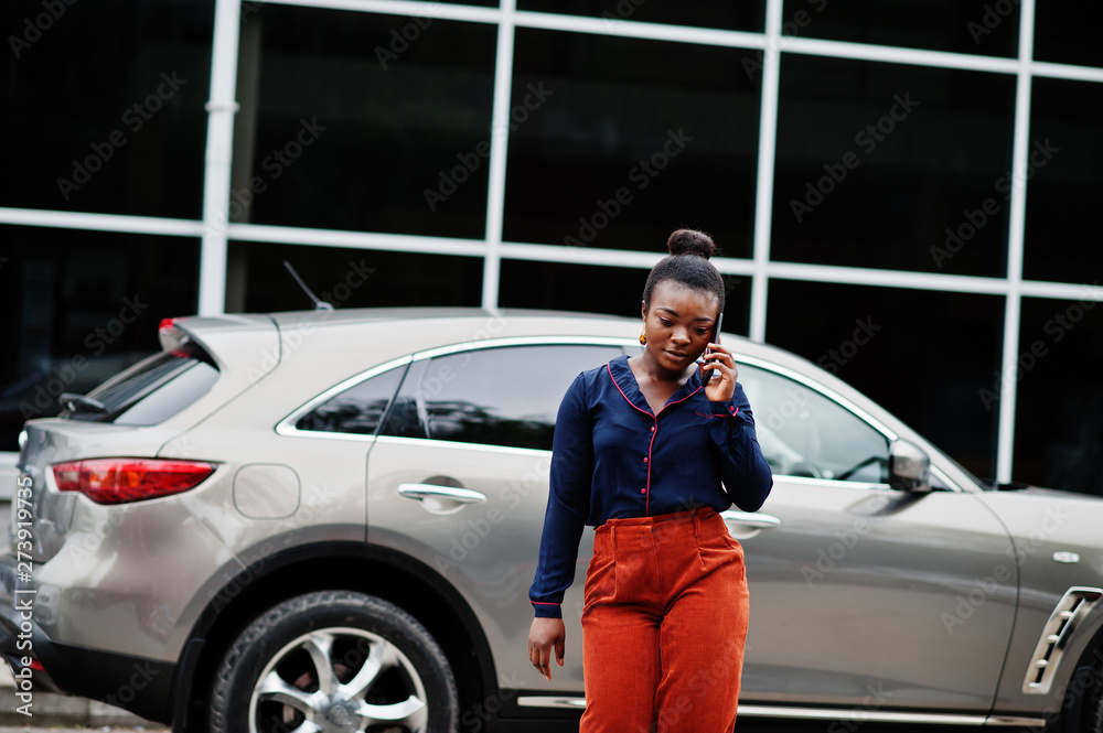 Rich business african woman in orange pants and blue shirt posed against silver suv car and speak on mobile phone.