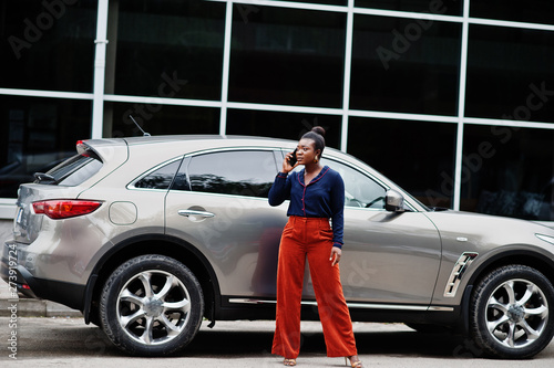Rich business african woman in orange pants and blue shirt posed against silver suv car and speak on mobile phone. © AS Photo Family