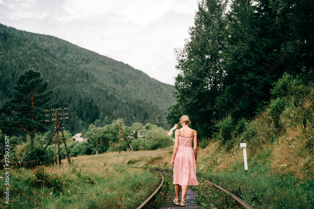 Naklejka premium Young girl walking on the railway. Woman in pink dress walking on railroad tracks.