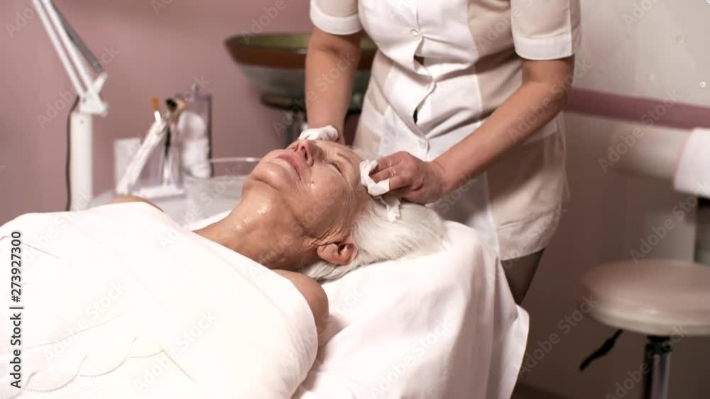 Tilting up medium shot of senior Caucasian lady with silver hair relaxing on couch with closed eyes in spa, and middle-aged brunette female beautician gently cleansing her face with moist wipes Tilting up medium shot of senior Caucasian lady with silver hair relaxing on couch with closed eyes in spa, and middle-aged brunette female beautician gently cleansing her face with moist wipes
