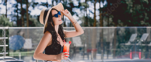 Fototapeta Naklejka Na Ścianę i Meble -  beautiful brunette young woman in swimsuit, straw hat and sunglasses smiling while holding cocktail, panoramic shot