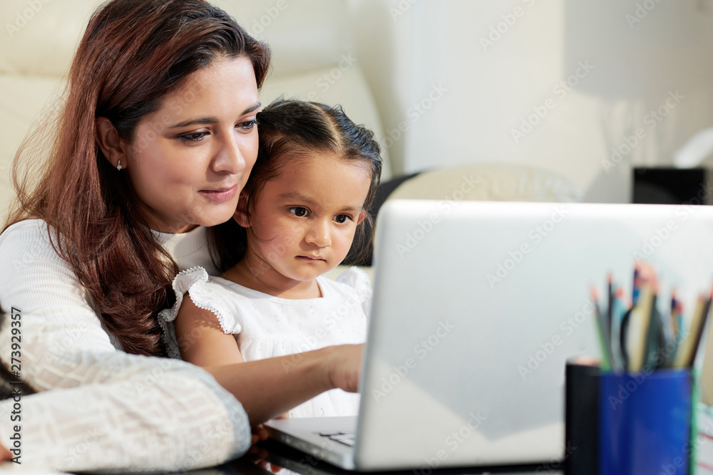 Young mother sitting at the table together with her little daughter and they typing on laptop computer at home Young mother sitting at the table together with her little daughter and they typing on laptop computer at home