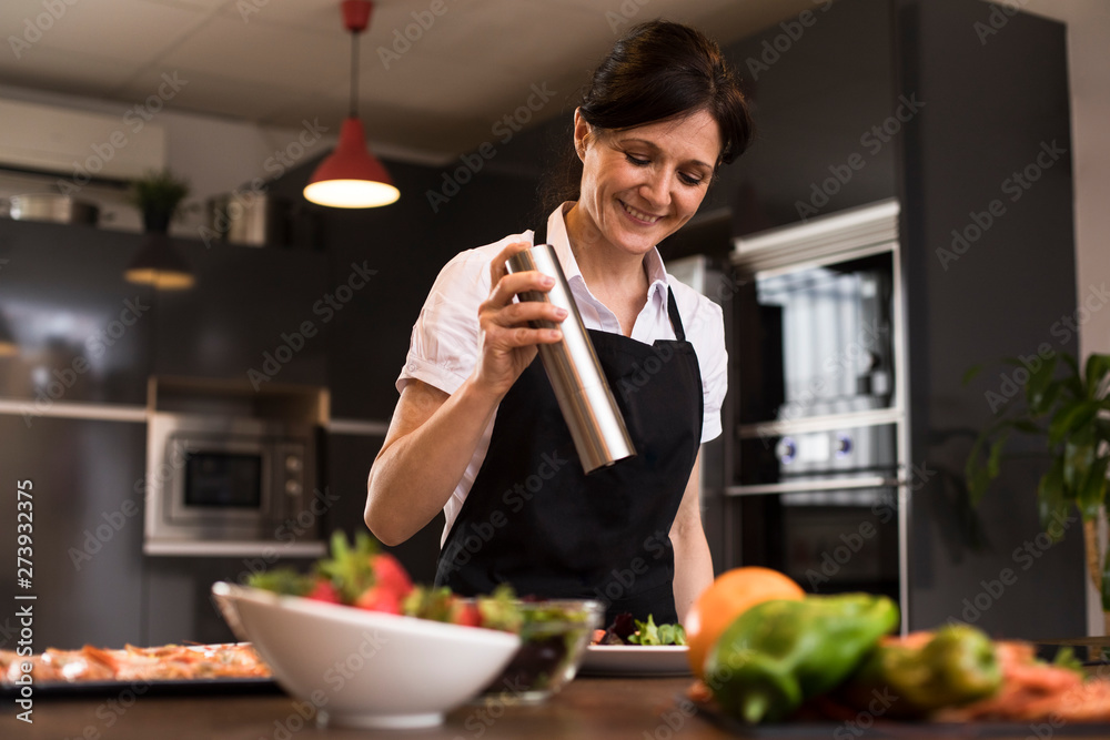Smiling woman cooking in kitchen pouring pepper on a dish Smiling woman cooking in kitchen pouring pepper on a dish