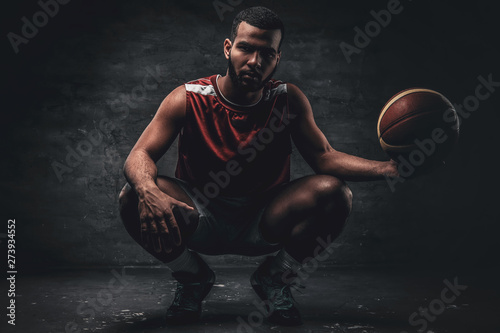 Full body portrait of a black basketball player sits on a floor and holds basket ball.