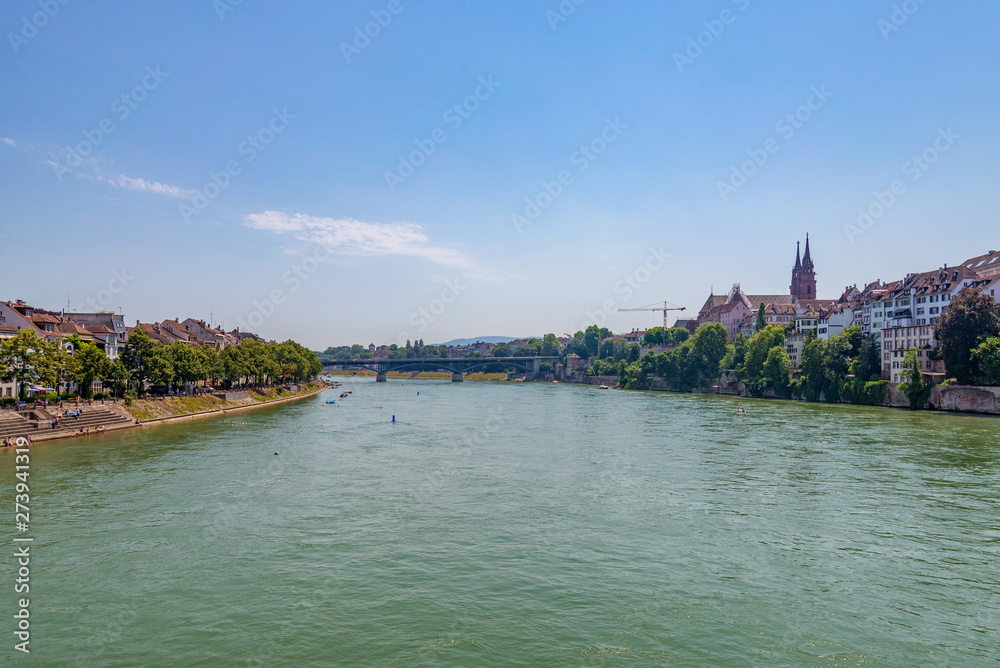 Outdoor sunny scenery of promenade and bridge along riverside, Basel's cityscape, Münster Cathedral and people float and swim, from the bridge cross Rhine river in Basel, Switzerland in summer season.