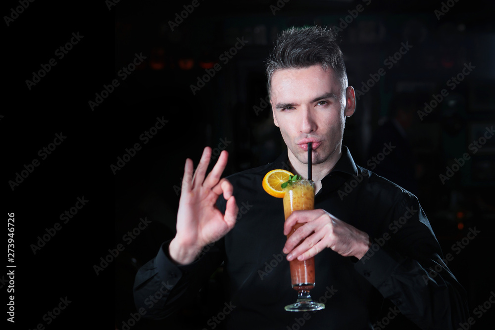 Fototapeta premium A handsome guy in a black shirt is holding a cocktail of orange juice in his hand and showing a hand gesture. Portrait of a guy of European appearance on a black background. Copy space.