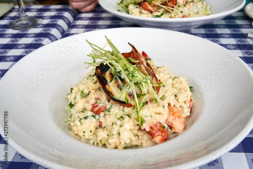 close up of lobster risotto in a white bowl topped with a half a grilled lobster tail sitting on a table with a blue and white checkered tablecloth