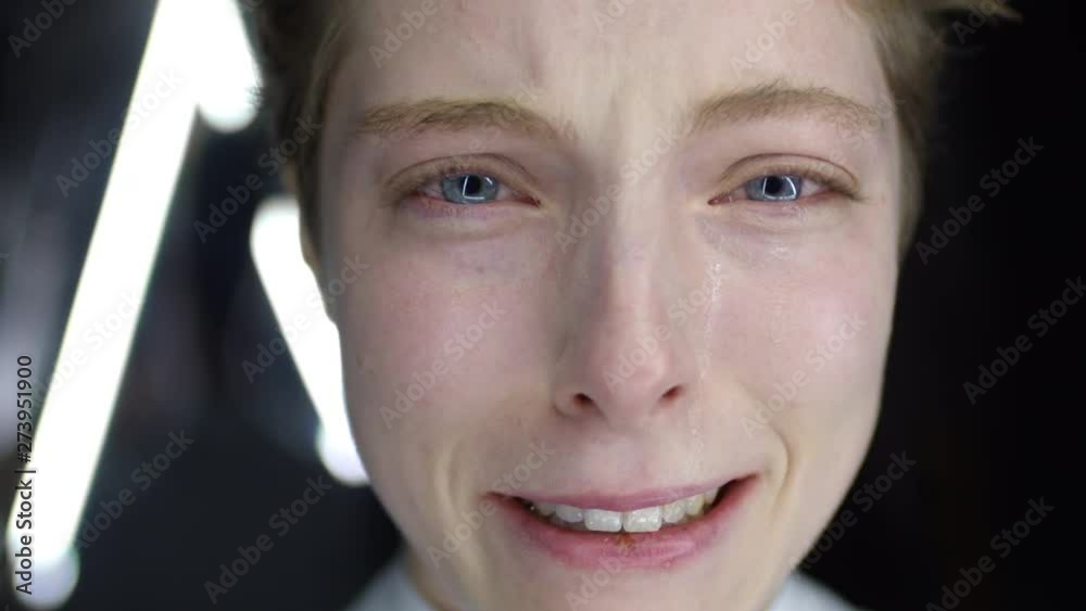 Macro of Caucasian young female face looking down then looking at camera and crying on dark background with triangular neon lamps Macro of Caucasian young female face looking down then looking at camera and crying on dark background with triangular neon lamps