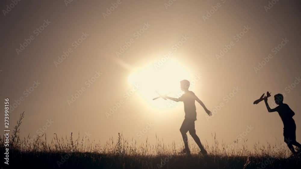 Two boys with a plane in their hands run across a green field in the rays of the suns at sunset. Children's dream of travel. Sports games for children Two boys with a plane in their hands run across a green field in the rays of the suns at sunset. Children's dream of travel. Sports games for children