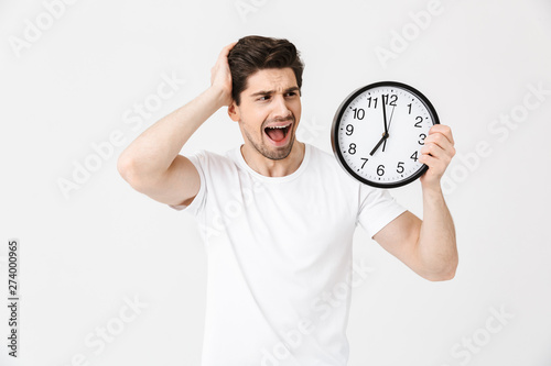 Screaming confused young man posing isolated over white wall background holding clock.