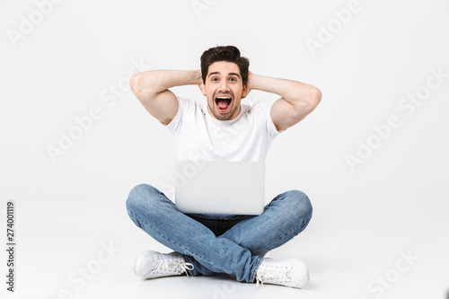 Happy excited young man posing isolated over white wall using laptop computer sitting on floor screaming. © Drobot Dean