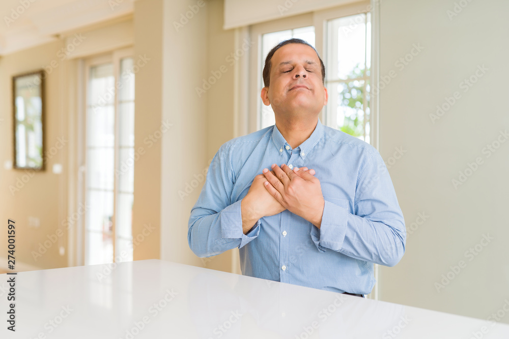 Middle age man sitting at home smiling with hands on chest with closed eyes and grateful gesture on face. Health concept.