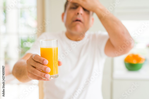 Middle age man drinking a glass of orange juice at home stressed with hand on head, shocked with shame and surprise face, angry and frustrated. Fear and upset for mistake.