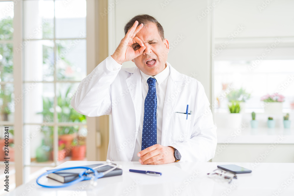 Middle age doctor man wearing medical coat at the clinic doing ok gesture with hand smiling, eye looking through fingers with happy face.
