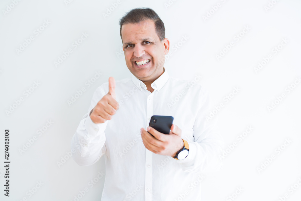 Middle age man using smartphone over white wall happy with big smile doing ok sign, thumb up with fingers, excellent sign