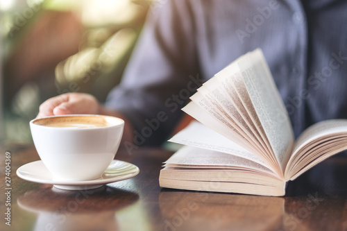 Closeup image of a woman holding and reading a book while drinking coffee on wooden table