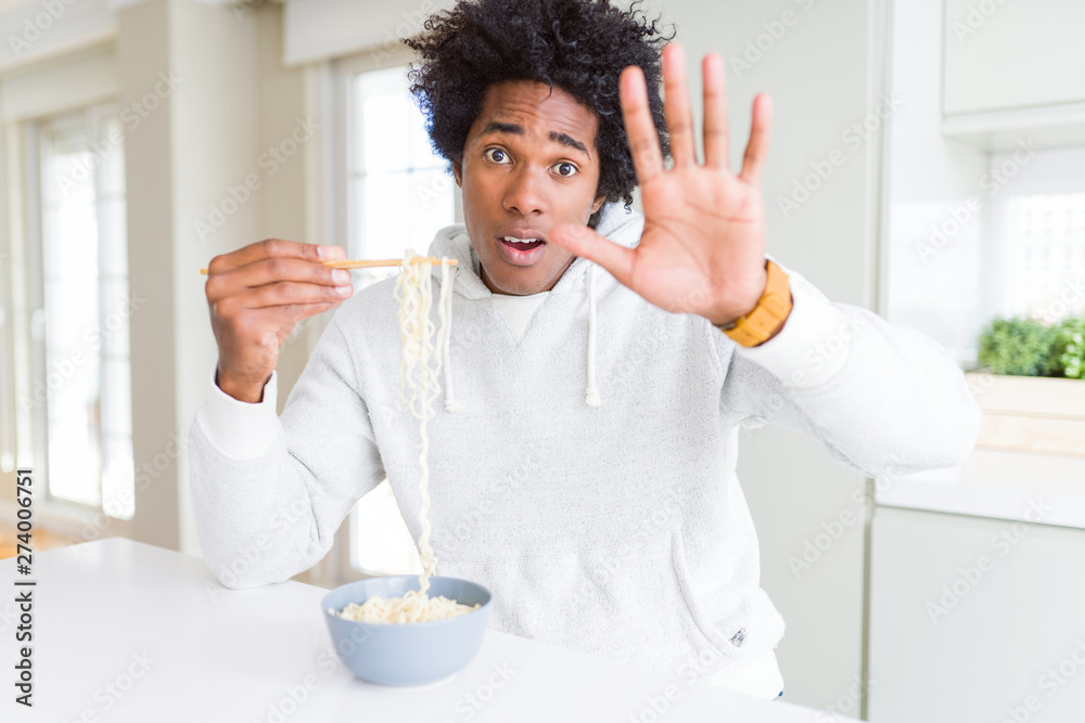 African American man eating asian noodles using chopsticks at home with open hand doing stop sign with serious and confident expression, defense gesture African American man eating asian noodles using chopsticks at home with open hand doing stop sign with serious and confident expression, defense gesture