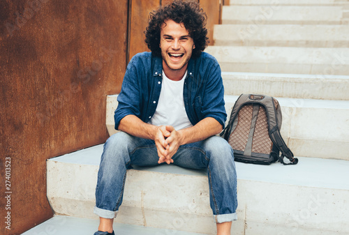 Outdoor image of handsome smiling young man student with backpack stitting at concrete stairs on the city street. Positive student male wearing casual outfit, relaxing outdoors. People and lifestyle photo