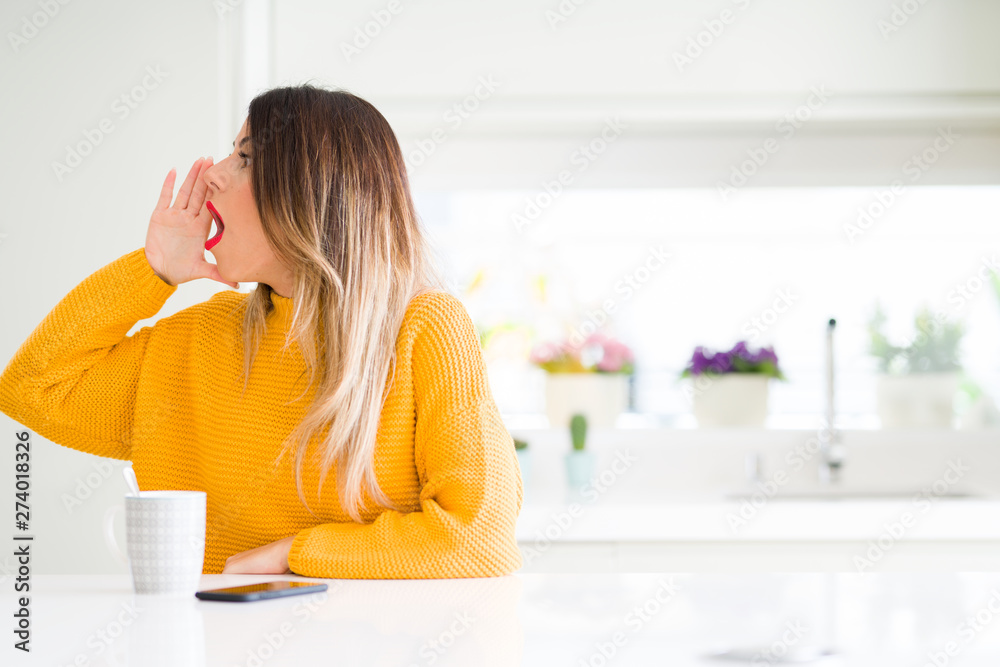 Young beautiful woman drinking a cup of coffee at home shouting and screaming loud to side with hand on mouth. Communication concept. Young beautiful woman drinking a cup of coffee at home shouting and screaming loud to side with hand on mouth. Communication concept.