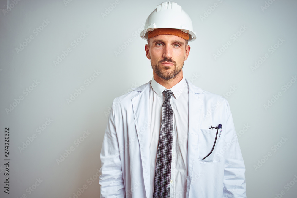 Young handsome engineer man wearing safety helmet over isolated background with serious expression on face. Simple and natural looking at the camera.