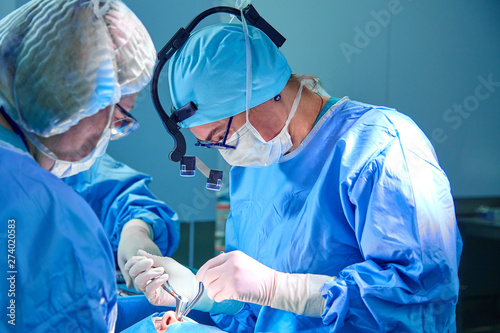 Surgeon and his assistant performing cosmetic surgery in hospital operating room. Surgeon in mask wearing loupes during medical procadure.