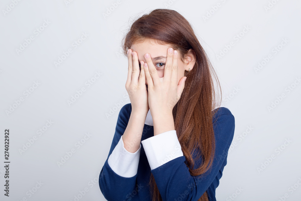 Red haired girl looks through her fingers covering her face with her hands, standing against a gray background. Schoolgirl. Red haired girl looks through her fingers covering her face with her hands, standing against a gray background. Schoolgirl.