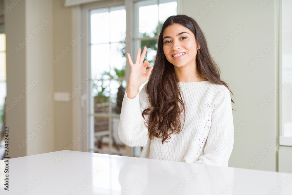 Young beautiful woman at home on white table smiling positive doing ok sign with hand and fingers. Successful expression.