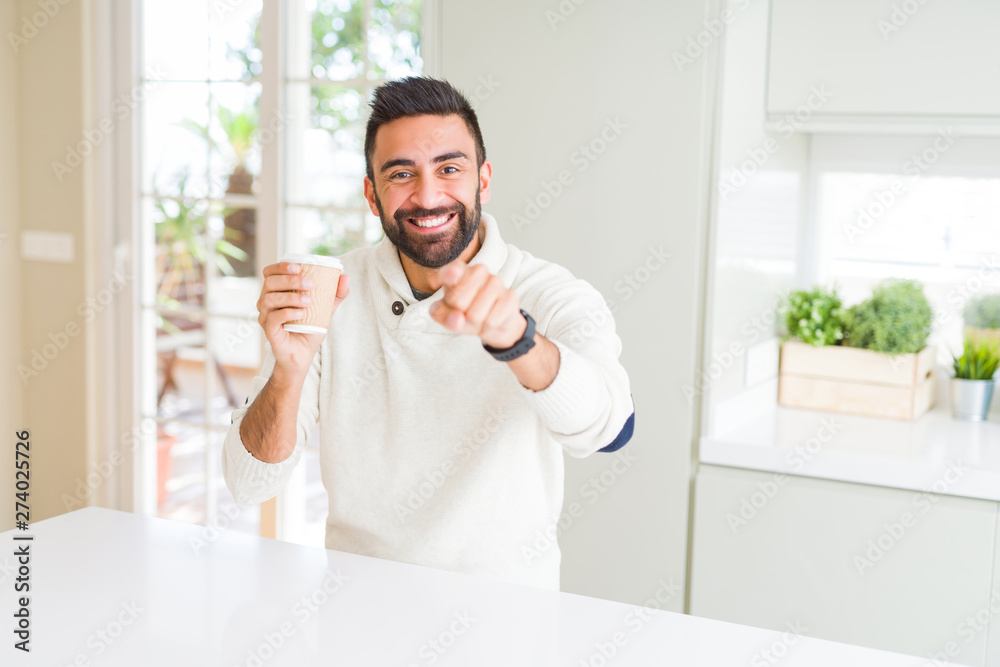 Handsome hispanic man drinking a coffee in a paper cup pointing with finger to the camera and to you, hand sign, positive and confident gesture from the front