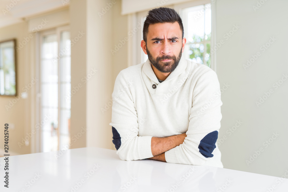 Handsome hispanic man wearing casual white sweater at home skeptic and nervous, disapproving expression on face with crossed arms. Negative person.