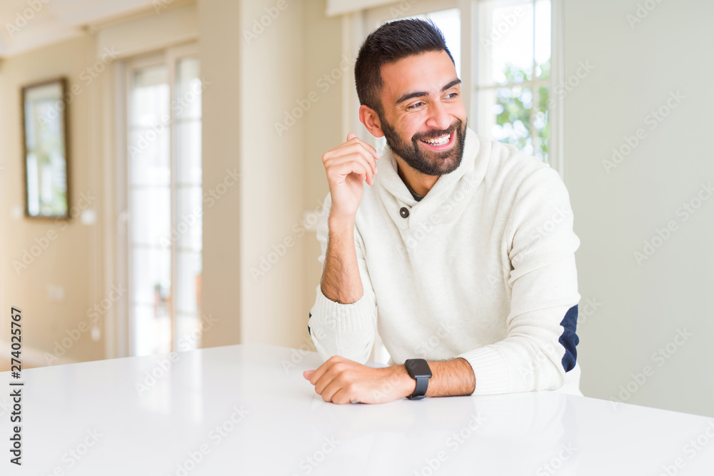 Handsome hispanic man wearing casual white sweater at home looking away to side with smile on face, natural expression. Laughing confident.