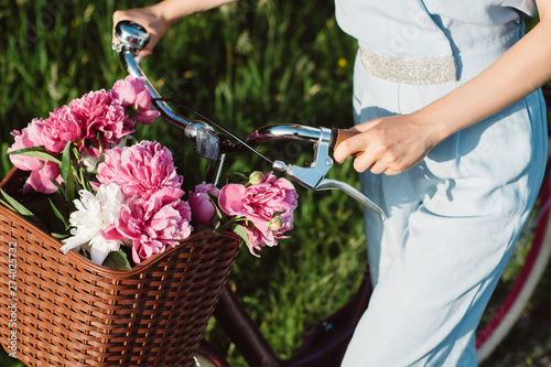 Fototapeta Naklejka Na Ścianę i Meble -  Close up shot of female hands, she holds a bicycle with a basket of red and white peonies flowers. Active vacation concept.