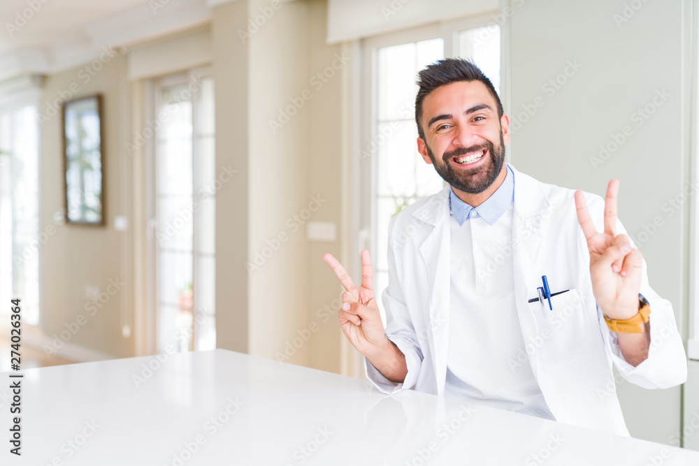 Handsome hispanic doctor or therapist man wearing medical coat at the clinic smiling looking to the camera showing fingers doing victory sign. Number two. Handsome hispanic doctor or therapist man wearing medical coat at the clinic smiling looking to the camera showing fingers doing victory sign. Number two.
