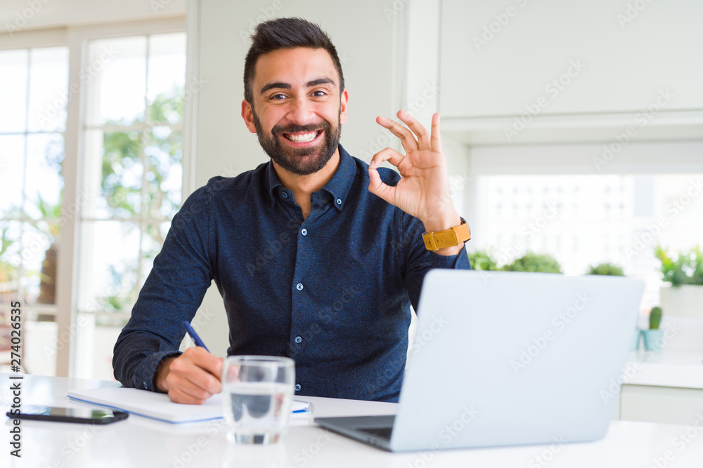 Fotografie Handsome hispanic man working using computer and writing on a paper doing ok sig