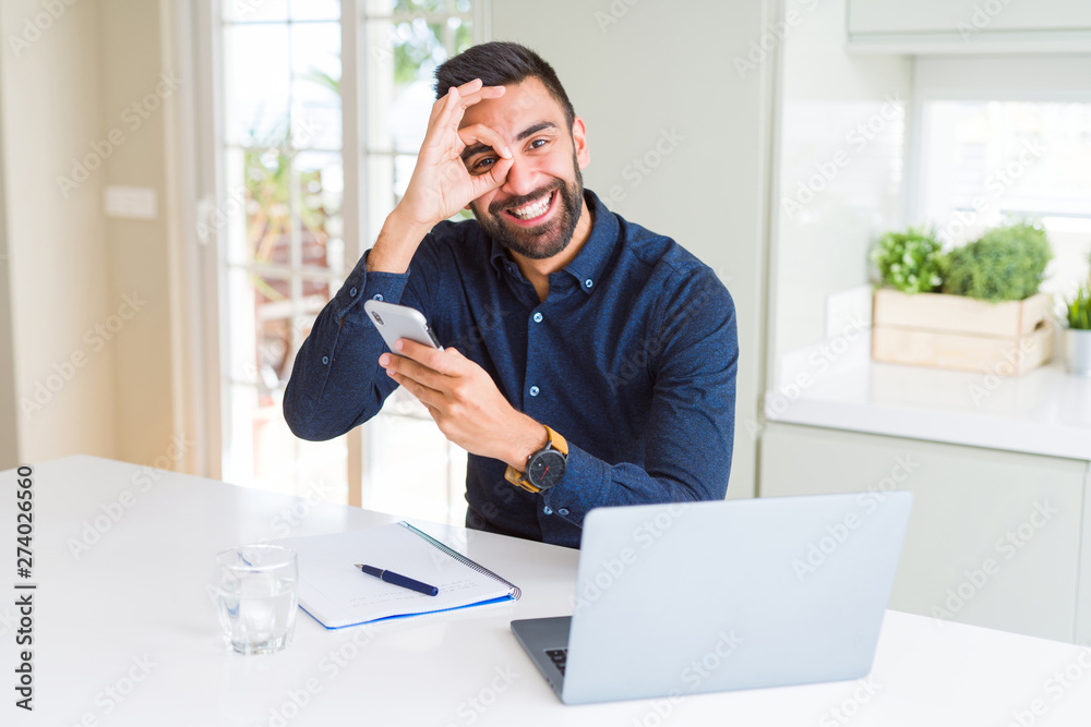 Handsome hispanic business man using smartphone and laptop at the office with happy face smiling doing ok sign with hand on eye looking through fingers