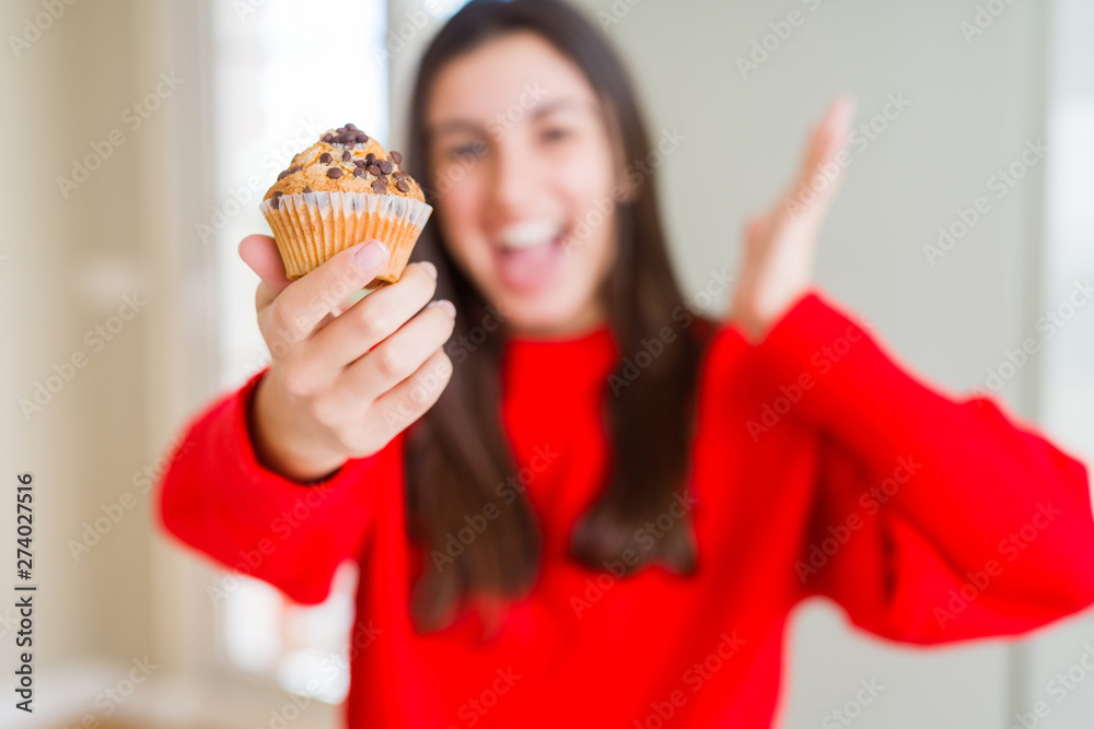 Beautiful young woman eating chocolate chips muffin very happy and excited, winner expression celebrating victory screaming with big smile and raised hands