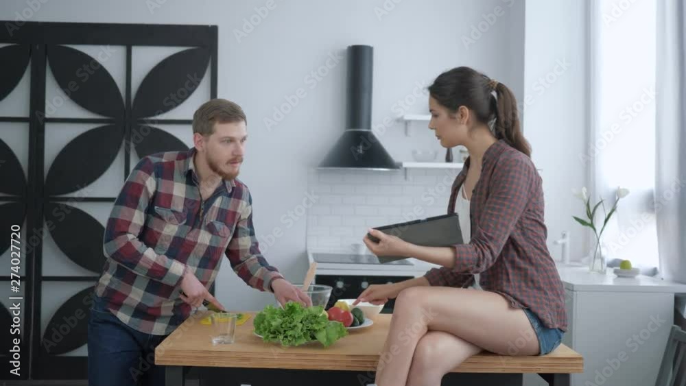 healthy eating, attractive male prepares useful vegetarian food on dinner and female sitting on kitchen table with tablet in hands and and advice to cooking at cuisine healthy eating, attractive male prepares useful vegetarian food on dinner and female sitting on kitchen table with tablet in hands and and advice to cooking at cuisine
