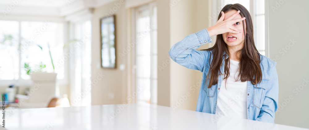 Wide angle picture of beautiful young woman sitting on white table at home peeking in shock covering face and eyes with hand, looking through fingers with embarrassed expression.