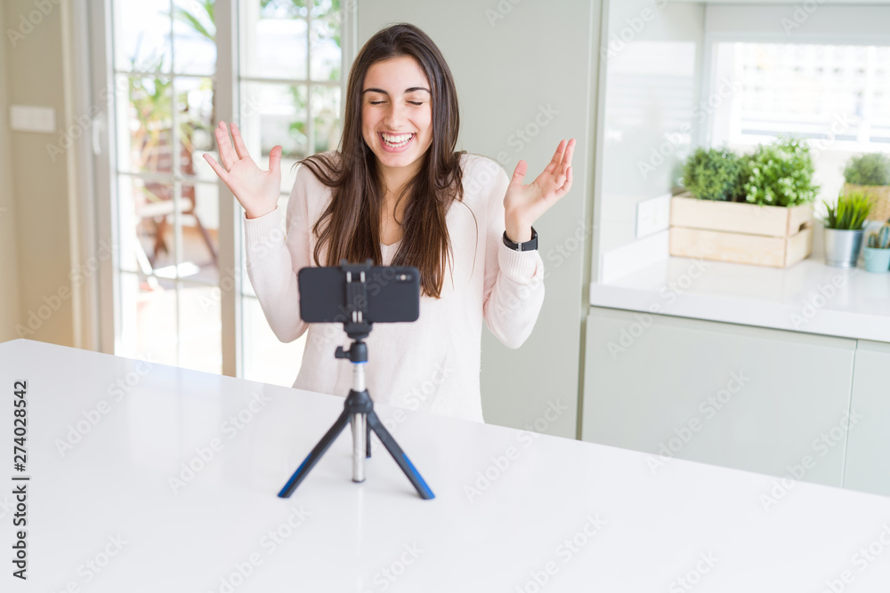Beautiful young woman recording selfie video with smartphone webcam celebrating mad and crazy for success with arms raised and closed eyes screaming excited. Winner concept Beautiful young woman recording selfie video with smartphone webcam celebrating mad and crazy for success with arms raised and closed eyes screaming excited. Winner concept