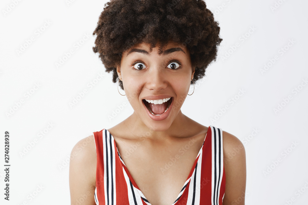 I am amazed, it is fantastic. Portrait of thrilled and charmed african-american woman with afro haircut, gasping from happiness, smiling broadly and gazing amused at camera, listening carefully