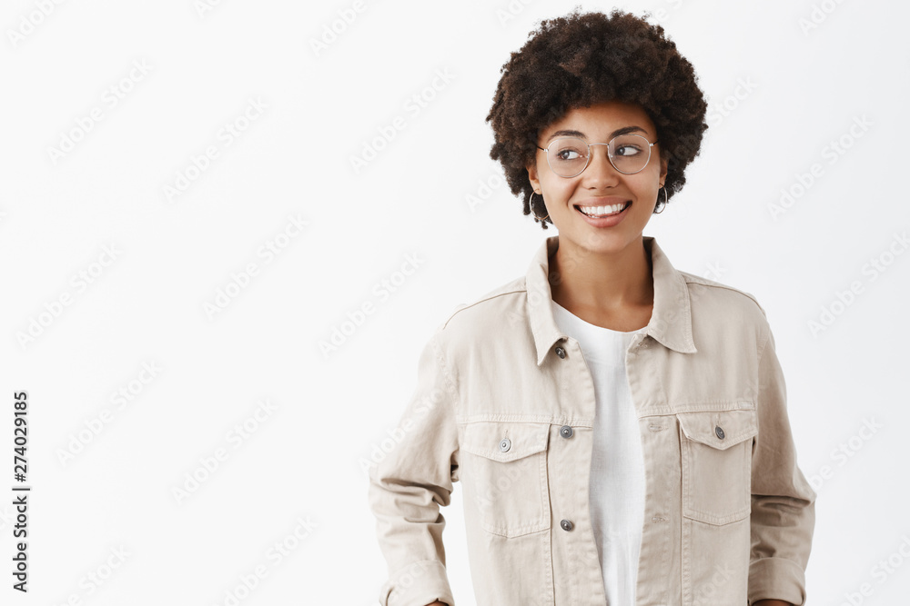 Studio shot of creative confident and carefree dark-skinned female freelancer in beige shirt and glasses holding hands in pockets gazing left joyfully, spending great time over gray wall Studio shot of creative confident and carefree dark-skinned female freelancer in beige shirt and glasses holding hands in pockets gazing left joyfully, spending great time over gray wall