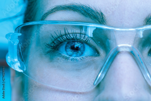 A close up view on the eyes of a pretty young Caucasian lady, working as a technician inside a laboratory, she wears protective goggles, cool blue filter gives futuristic atmosphere. photo