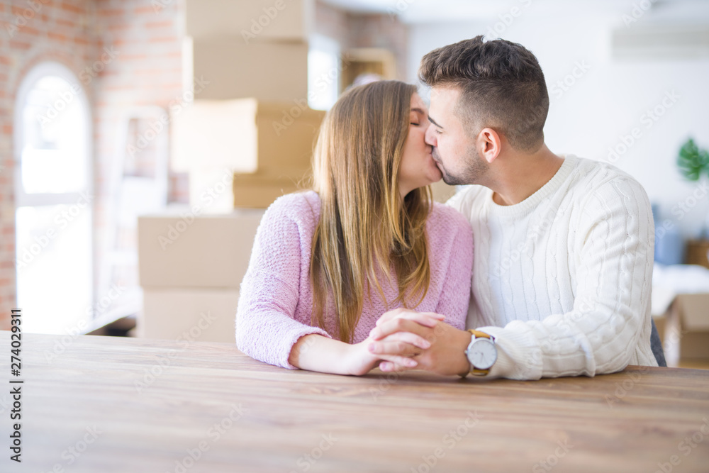 Young beautiful couple sitting on the table at home, hugging in love very happy for moving to new home with cardboard boxes behind them Young beautiful couple sitting on the table at home, hugging in love very happy for moving to new home with cardboard boxes behind them