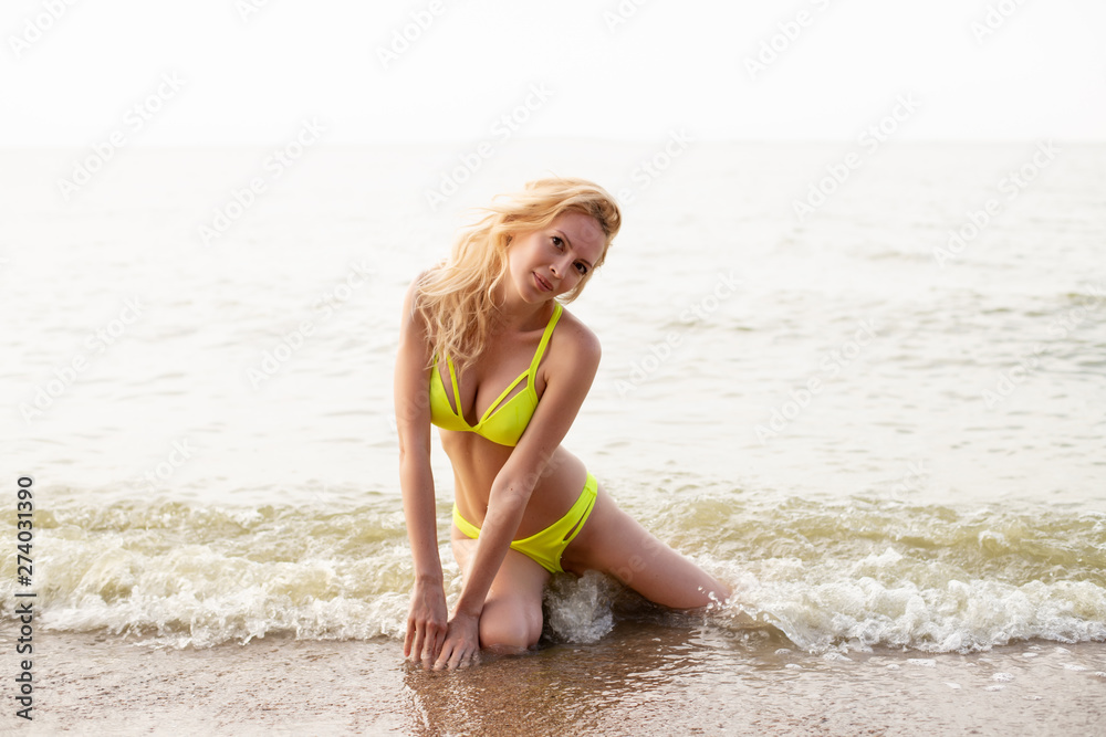 Naklejka premium Beautiful young girl with a slim perfect sexy body sits in the water on a sandy beach by the sea