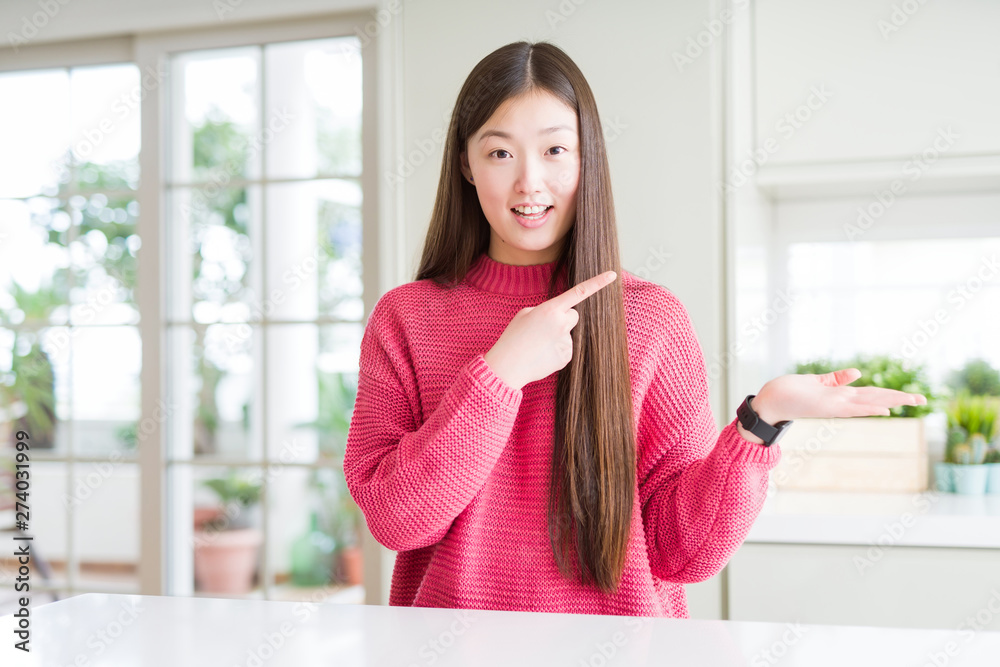 Beautiful Asian woman wearing pink sweater on white table amazed and smiling to the camera while presenting with hand and pointing with finger.