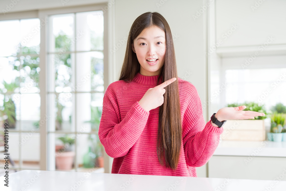 Beautiful Asian woman wearing pink sweater on white table amazed and smiling to the camera while presenting with hand and pointing with finger. Beautiful Asian woman wearing pink sweater on white table amazed and smiling to the camera while presenting with hand and pointing with finger.