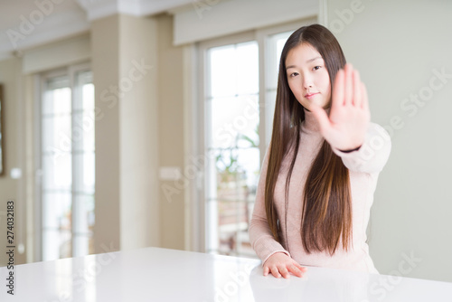 Beautiful Asian woman wearing casual sweater on white table doing stop sing with palm of the hand. Warning expression with negative and serious gesture on the face.