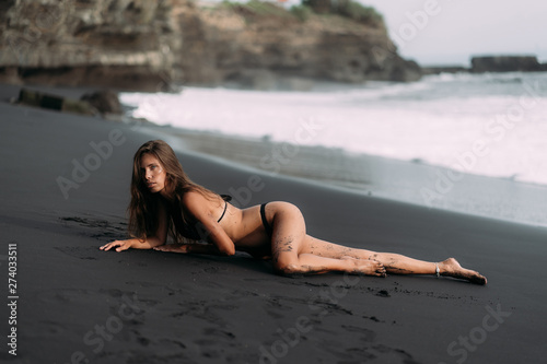 Young sexy girl in black swimwear resting on black volcanic sand beach