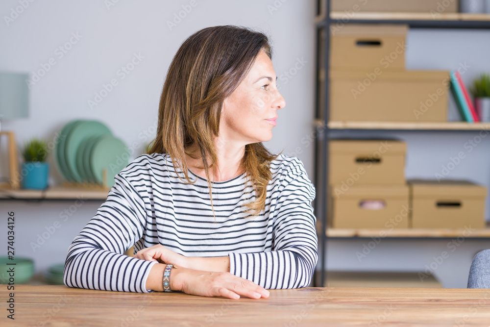 Middle age senior woman sitting at the table at home looking to side, relax profile pose with natural face with confident smile. Middle age senior woman sitting at the table at home looking to side, relax profile pose with natural face with confident smile.