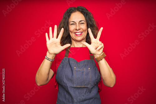 Middle age senior woman wearing apron uniform over red isolated background showing and pointing up with fingers number eight while smiling confident and happy.